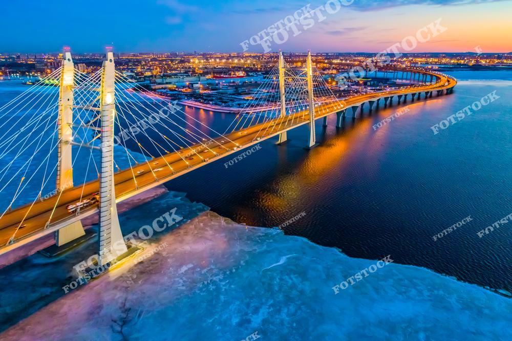 На фотографии изображён Большой Обуховский мост — единственный вантовый мост через Неву в Санкт-Петербурге. Мост является частью Западного скоростного диаметра (ЗСД), соединяющего юг и север города. Снимок сделан в вечернее время, когда городские огни отражаются в тёмных водах реки. Замёрзшая Нева подчёркивает зимнюю атмосферу мегаполиса. Это один из самых технологичных и протяжённых мостов города.